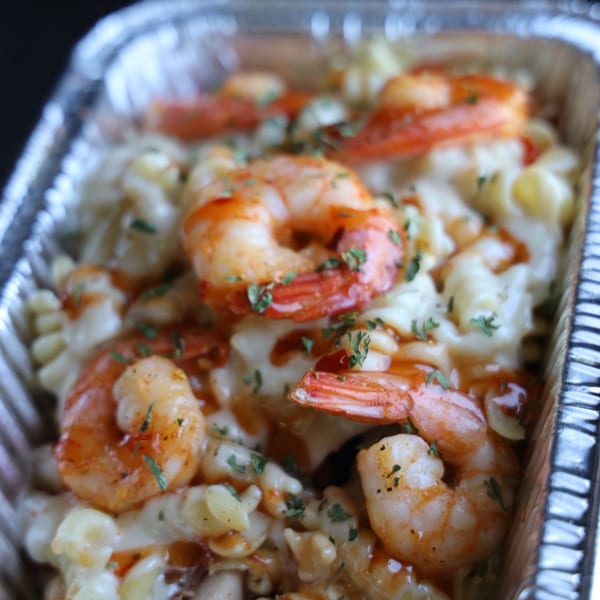 Shrimp mac and cheese with parsley and hot sauce in a foil takeout container.