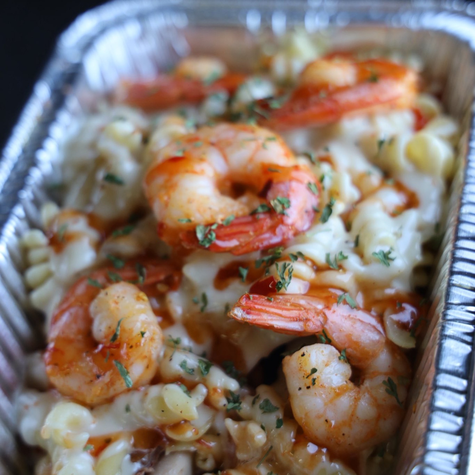 Shrimp mac and cheese with parsley and hot sauce in a foil takeout container.